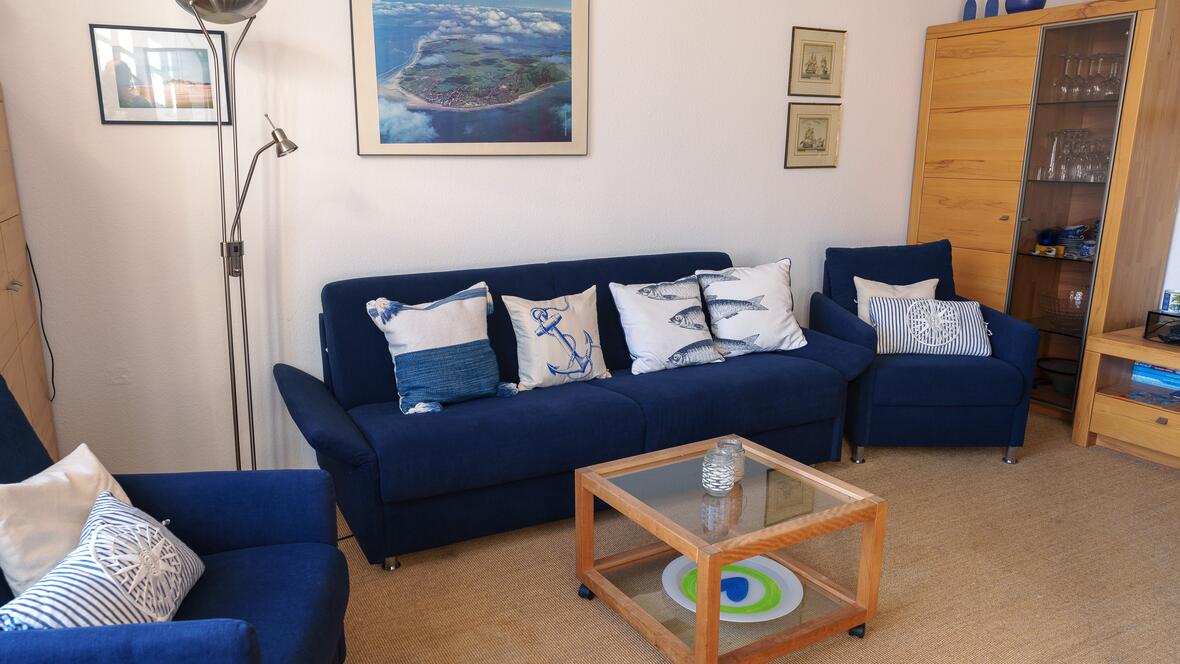 Das helle Wohnzimmer der Ferienwohnung Charlotte auf Borkum bietet eine gemütliche Sitzecke mit blauem Sofa, maritimer Dekoration und wohnlicher Ausstattung für einen entspannten Urlaub auf der Nordsee Insel.